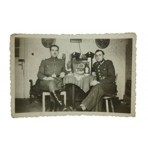 II RP Dwóch siedzących przy stole żołnierzy, w tle radio i dwie rogatywki na nim